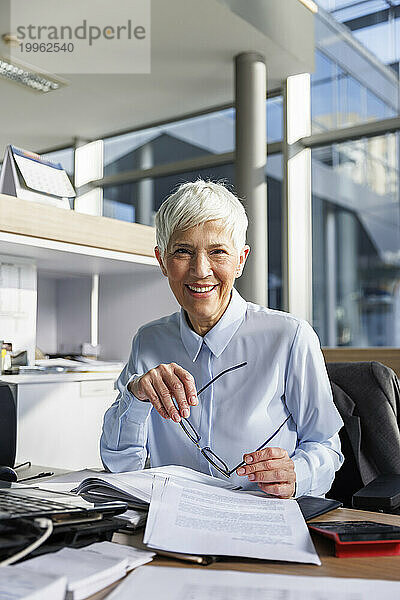 Glückliche ältere Geschäftsfrau mit Brille sitzt am Schreibtisch im Büro