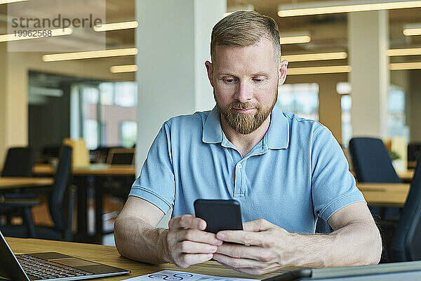 Geschäftsmann benutzt Smartphone neben Laptop am Schreibtisch im Büro