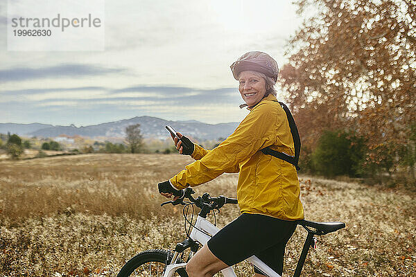 Glückliche Frau sitzt auf dem Mountainbike mit Smartphone im Feld