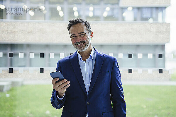 Glücklicher Geschäftsmann hält Smartphone vor Glas im Büro