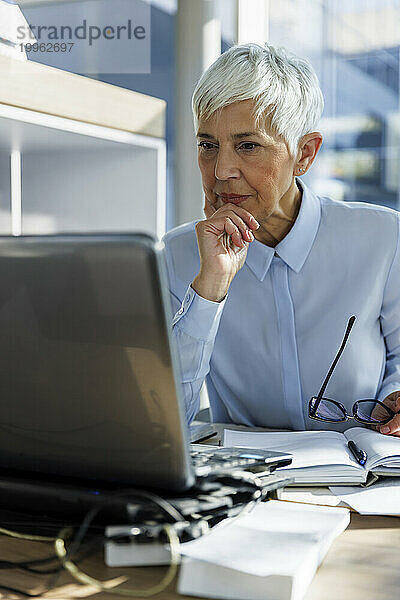 Nachdenkliche Geschäftsfrau mit der Hand am Kinn und Blick auf den Laptop im Büro