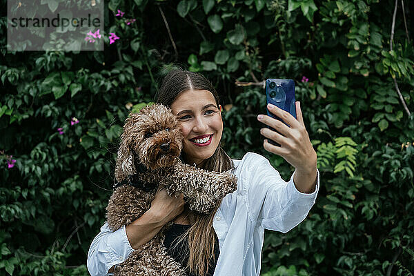 Glückliche Frau macht Selfie mit Pudelhund vor Pflanzen