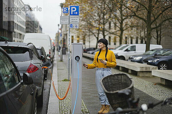 Frau mit Smartphone steht an der Ladestation für Elektrofahrzeuge