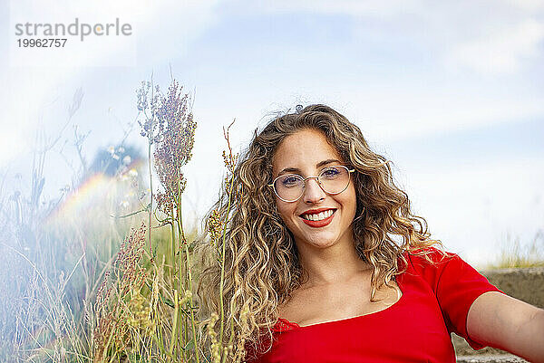 Smiling young woman with curly hair wearing eyeglasses near plants