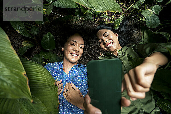 Lächelnde Frau macht Selfie auf Smartphone mit Freundin  die inmitten von Pflanzen liegt
