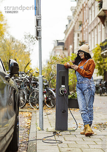 Frau benutzt Smartphone und lädt Elektroauto am Bahnhof auf