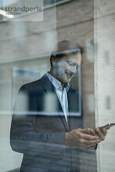 Lächelnder Geschäftsmann mit Mobiltelefon durch Glas gesehen