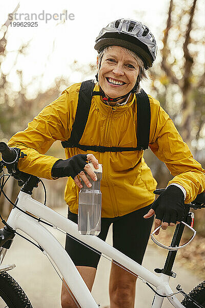 Glückliche Frau hält Wasserflasche in der Hand und stützt sich auf Mountainbike im Wald