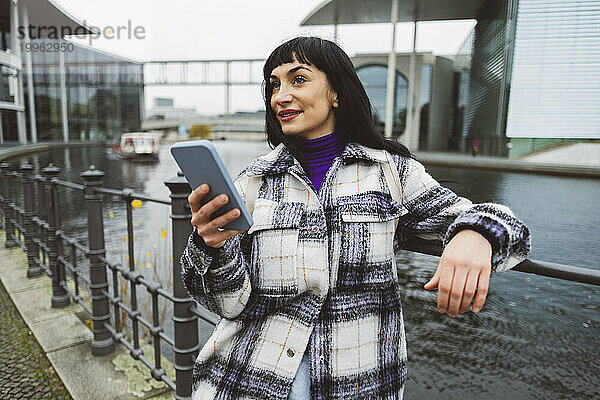 Nachdenkliche Frau mit Mobiltelefon steht vor dem Fluss