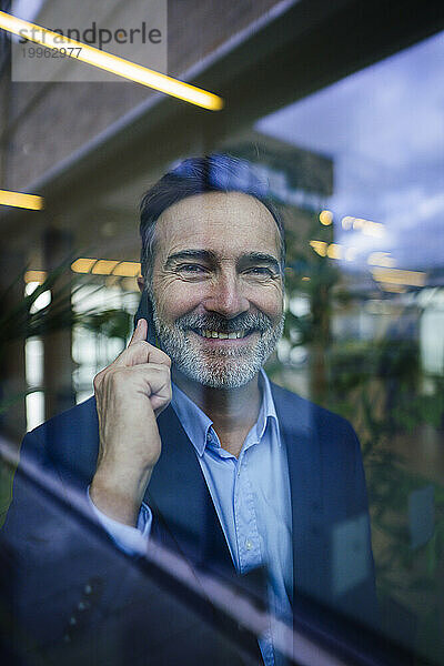 Glücklicher Geschäftsmann  der durch Glas auf dem Smartphone spricht