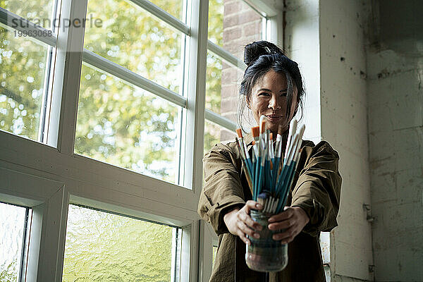 Smiling painter holding paintbrushes standing by window at workshop