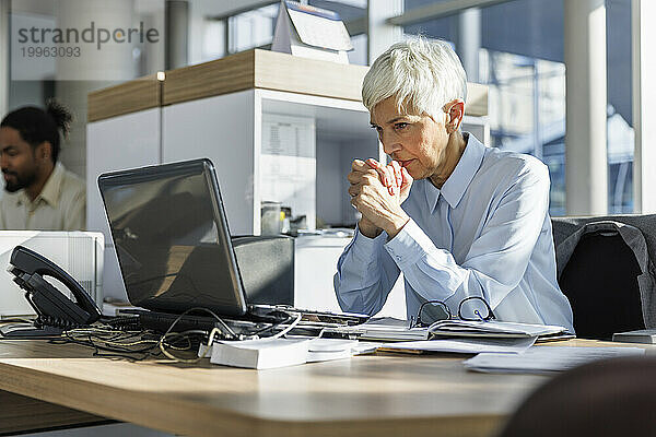 Besorgte Geschäftsfrau mit gefalteten Händen blickt im Büro auf den Laptop