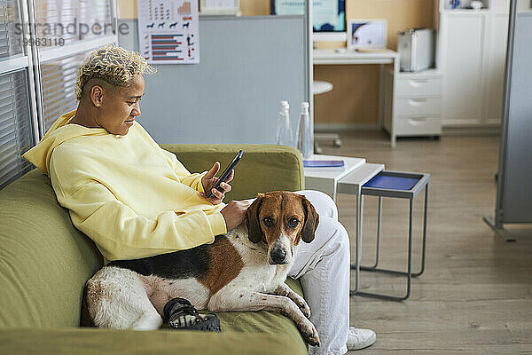 Frau benutzt Mobiltelefon und sitzt mit behindertem Hund auf Sofa in Tierklinik