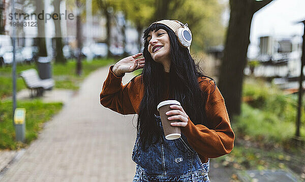 Glückliche schöne Frau hält eine Kaffeetasse in der Hand und genießt Musik über kabellose Kopfhörer auf dem Fußweg