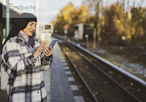 Lächelnde Frau  die auf dem Bahnsteig steht und ihr Mobiltelefon benutzt