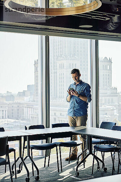 Reifer Geschäftsmann benutzt Smartphone am Fenster im Büro