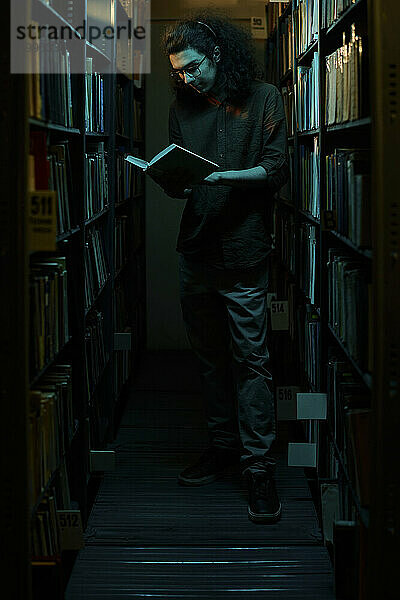 Student liest Buch und steht inmitten von Regalen in der Bibliothek
