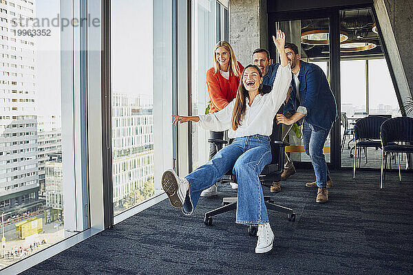 Fröhliche Kollegen drängen Geschäftsfrau  die auf einem Stuhl sitzt und Spaß im Büro hat