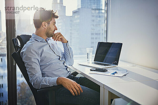 Nachdenklicher junger Geschäftsmann sitzt mit Laptop am Schreibtisch