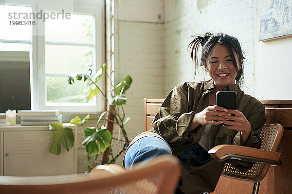 Lächelnder Künstler  der in der Werkstatt auf einem Stuhl sitzt und sein Smartphone benutzt