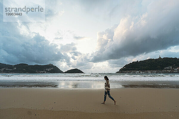 Junge Frau läuft unter bewölktem Himmel am Ufer der Playa de la Concha