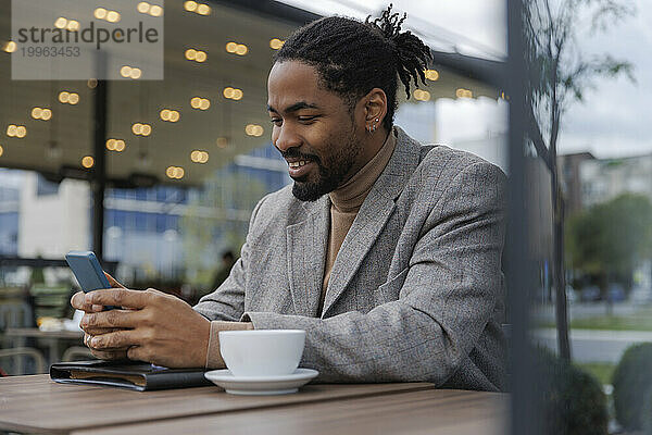 Lächelnder Geschäftsmann sitzt mit Kaffeetasse und benutzt Smartphone im Straßencafé