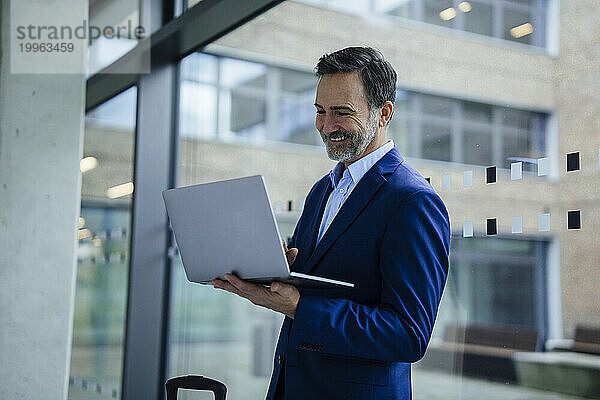 Lächelnder Geschäftsmann steht und benutzt Laptop neben Glas im Büro