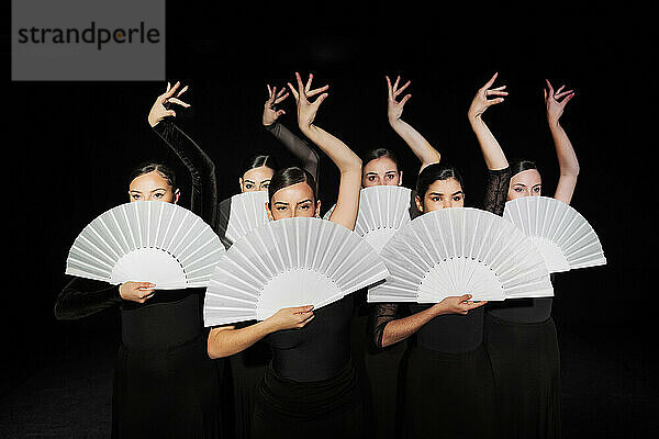 Group of flamenco dancers holding hand fans and dancing against black background