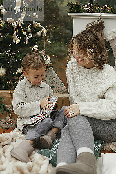 Lächelnde Mutter mit Sohn liest Buch neben dem Weihnachtsbaum zu Hause