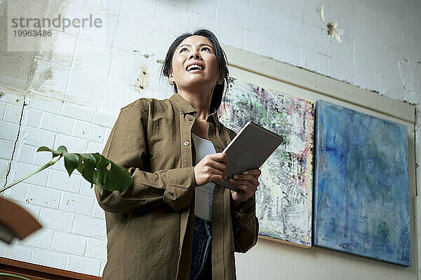 Lächelnde Frau hält Tablet-PC vor Gemälden im Kunstatelier