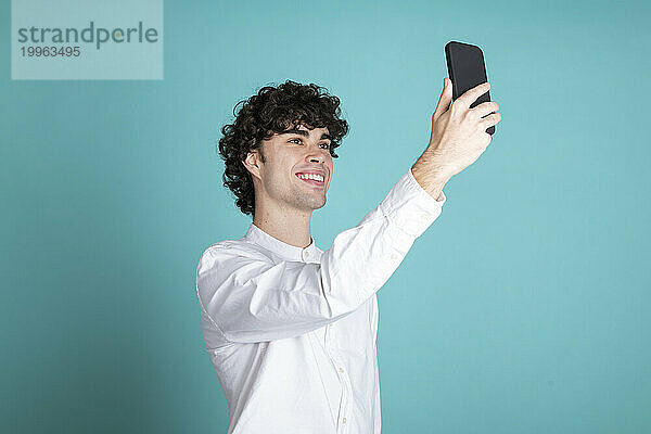 Lächelnder junger Mann macht ein Selfie mit dem Smartphone vor cyanfarbenem Hintergrund