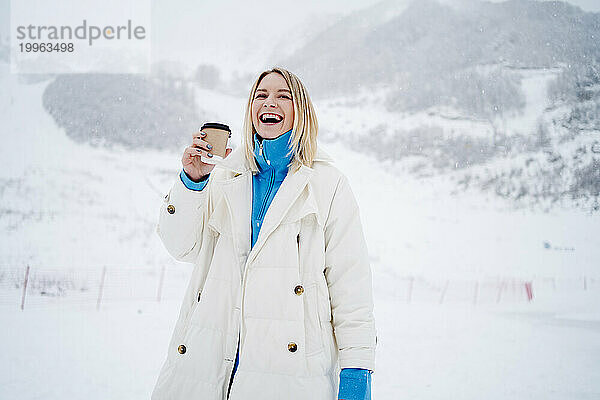Glückliche blonde Frau hält Kaffeetasse in der Hand und lacht auf einem schneebedeckten Berg