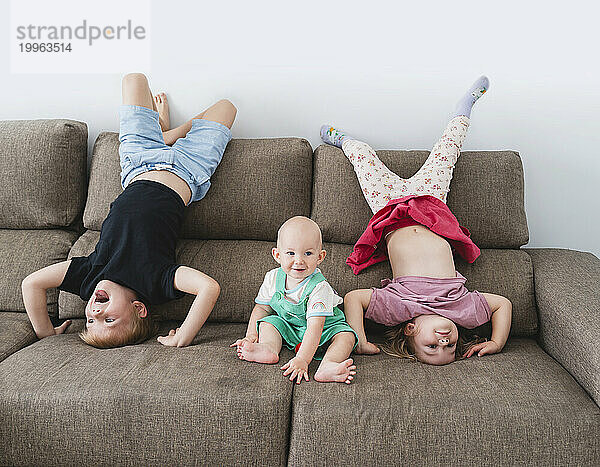 Glückliche Kinder machen Kopfstand neben ihrem kleinen Bruder auf dem heimischen Sofa