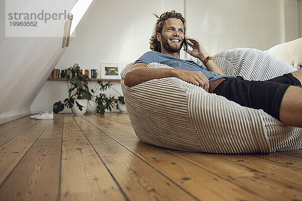 Smiling young man relaxing in beanbag at home talking on the phone