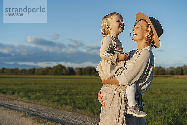 Glückliche schwangere Mutter trägt Tochter und lacht in der Nähe des Feldes bei Sonnenuntergang