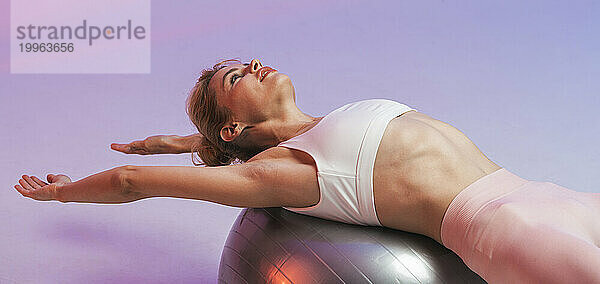 Frau trainiert auf einem Fitnessball mit ausgestreckten Armen vor zweifarbigem Hintergrund