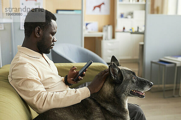 Man using smart phone and sitting with dog on sofa in clinic