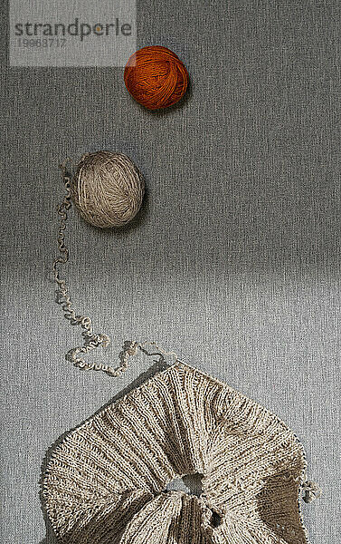Knitted sweater and balls of beige and terracotta yarn on table over gray background