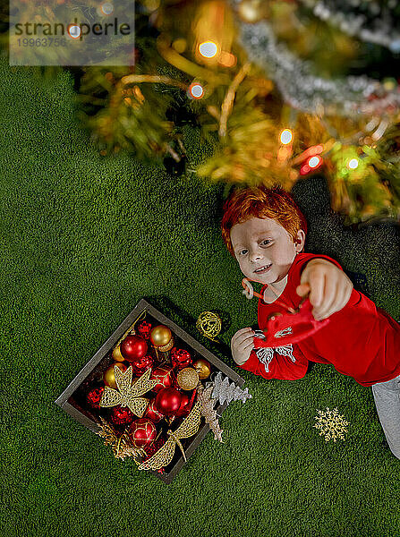Fröhlicher rothaariger Junge spielt mit Ornamenten am Weihnachtsbaum auf grünem Teppich