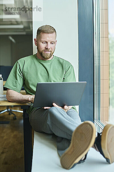 Lächelnder Geschäftsmann sitzt auf der Fensterbank und benutzt Laptop im Büro