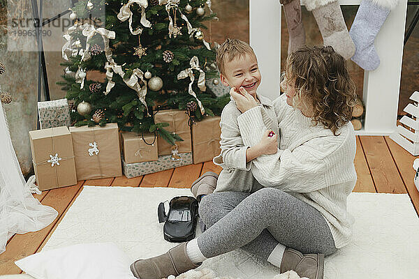 Glückliche Mutter umarmt ihren Sohn auf dem Teppich neben dem Weihnachtsbaum zu Hause