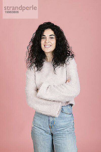 Smiling young woman standing with arms crossed against pink background