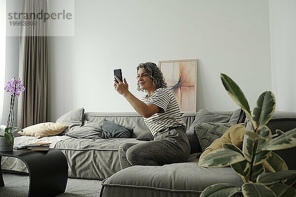 Reife Frau macht ein Selfie über ihr Smartphone und sitzt auf dem Sofa