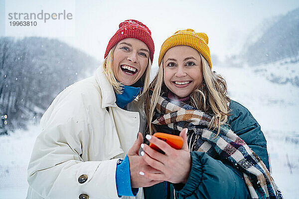 Lächelnde Freunde stehen mit Smartphone auf einem schneebedeckten Berg