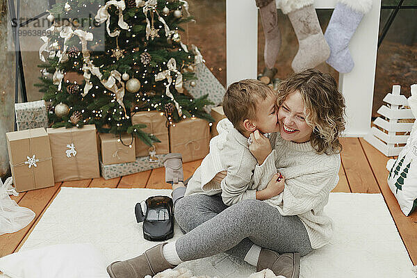 Junge küsst lächelnde Mutter  die zu Hause auf dem Teppich neben dem Weihnachtsbaum sitzt