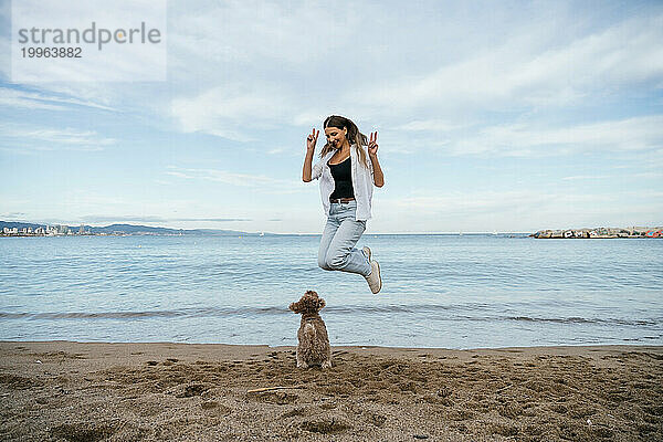 Verspielte junge Frau gestikuliert ein Friedenszeichen und springt neben Pudelhund am Strand