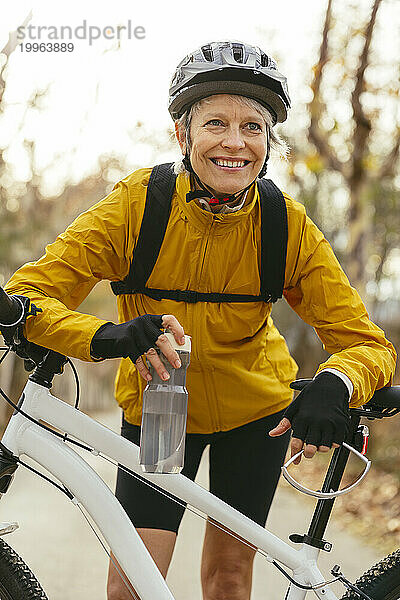 Lächelnde Frau hält Wasserflasche in der Hand und stützt sich auf Mountainbike im Wald