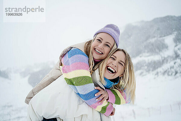 Glückliche blonde Frau gibt einer Freundin Huckepackfahrt auf einem schneebedeckten Berg unter freiem Himmel