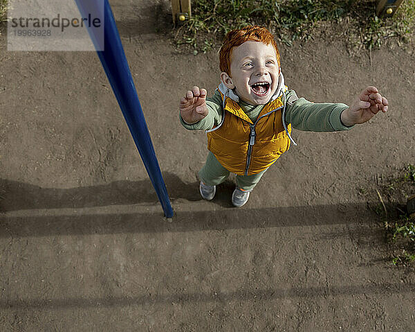 Fröhlicher rothaariger Junge steht mit erhobenen Armen auf dem Spielplatz