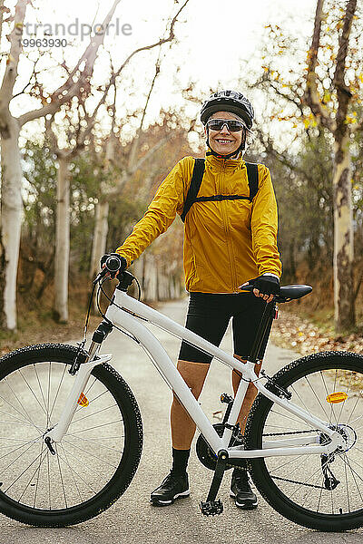 Lächelnde Frau steht mit Mountainbike auf der Straße im Wald